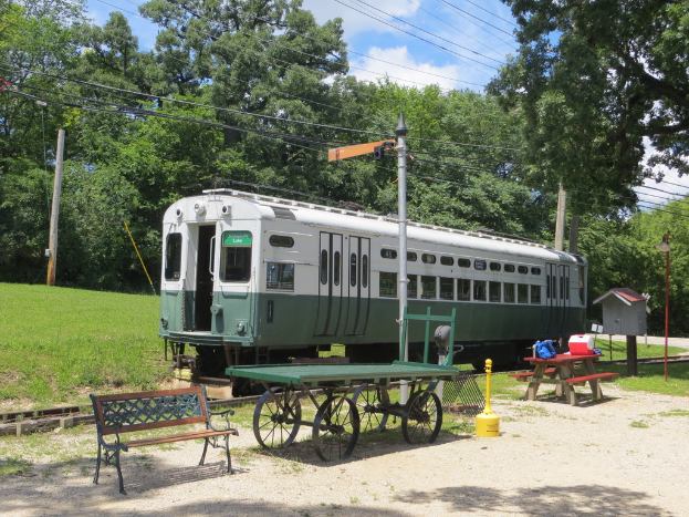 Ein Zug auf Schienen mit einer Bank, einem Tisch, einem Pfahl, Gras, Bäumen, Drähten und Himmel im Hintergrund.