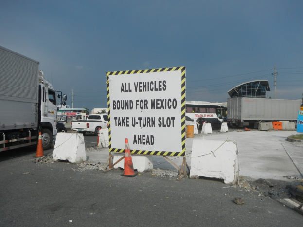 Lastwagen auf einem Parkplatz neben einem Schild mit der Aufschrift "Alle Fahrzeuge nach Mexiko" geparkt, mit verstreuten Verkehrskegeln, Brettern und Hintergrundgebäuden, Bäumen, Polen und einem klaren blauen Himmel.