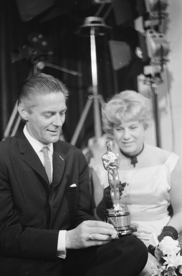 Ein Schwarz-Wei├č-Bild eines formell gekleideten Mannes mit einer Oscar-Statue und einer Frau mit einem Blumenstra├čus, die nebeneinander sitzen, mit beleuchteter Hintergrund.