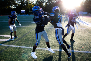 Junge Männer in Helmen und Fußballschuhen auf einem Feld mit Bäumen und blauem Himmel.