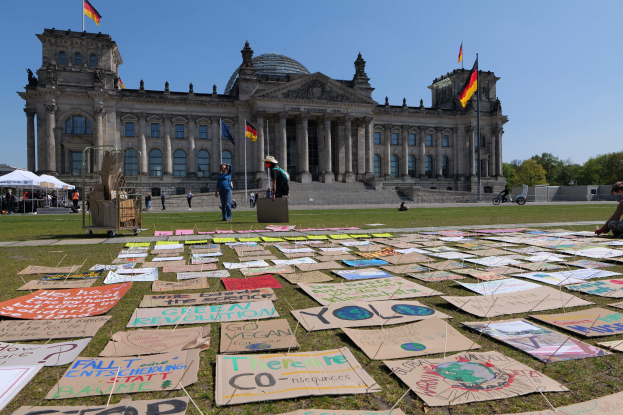 Große Gruppe von Menschen auf einem Rasenfeld vor dem Reichstaggebäude in Berlin, Deutschland, mit Zelten und verstreuten Gegenständen, Bäumen und einem klaren blauen Himmel im Hintergrund.