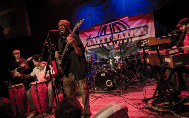 Eine Live-Band performt auf der Bühne mit Mikrofonen und Ständern, während das Publikum unten zuschaut; ein Banner mit Text ist im Hintergrund zu sehen.