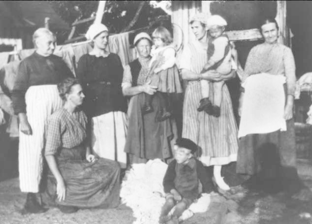 Eine Gruppe von Frauen steht vor einem Gebäude, mit einem Kind im Vordergrund auf dem Boden sitzend, auf einem Schwarz-Weiß-Bild, das den Holocaust zeigt, mit Wäsche auf einer Leine, einem Baum und einer Wand im Hintergrund.