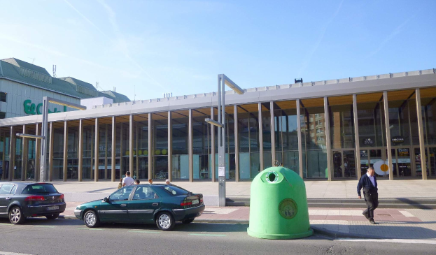 Grüner Müllcontainer auf einer Berliner Citystraße in der Nähe des Berliner Hauptbahnhofs, der Glaswände und Säulen hat, mit Fahrzeugen und Passanten im Hintergrund bei bewölktem Himmel.
