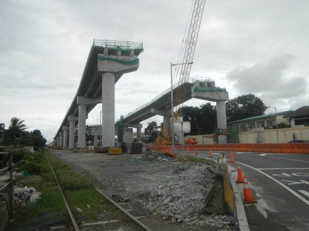 Baustelle mit einer Brücke im Hintergrund, eine Straße mit Absperrbaken auf der rechten Seite, Steine und Gras auf dem Boden, eine Bahnschiene auf der linken Seite, Bäume und Gebäude auf beiden Seiten und ein bewölkter Himmel.