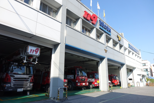 Feuerwache mit mehreren Feuerwehrautos vor der Tür, mit Fenstern, Säulen und Namensschildern, sowie Fahnen im Vordergrund und anderen Gebäuden im Hintergrund und einem klaren blauen Himmel.
