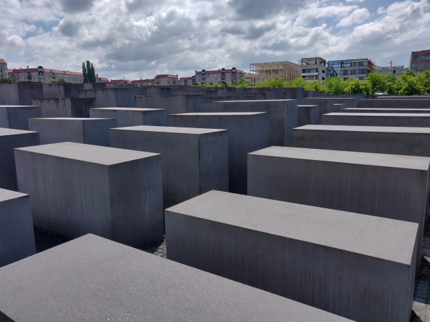 Betonschollen in einem Gittermuster auf dem Holocaust-Mahnmal für die ermordeten Juden Europas in Berlin, mit Bäumen und Gebäuden im Hintergrund unter einem bewölkten Himmel.