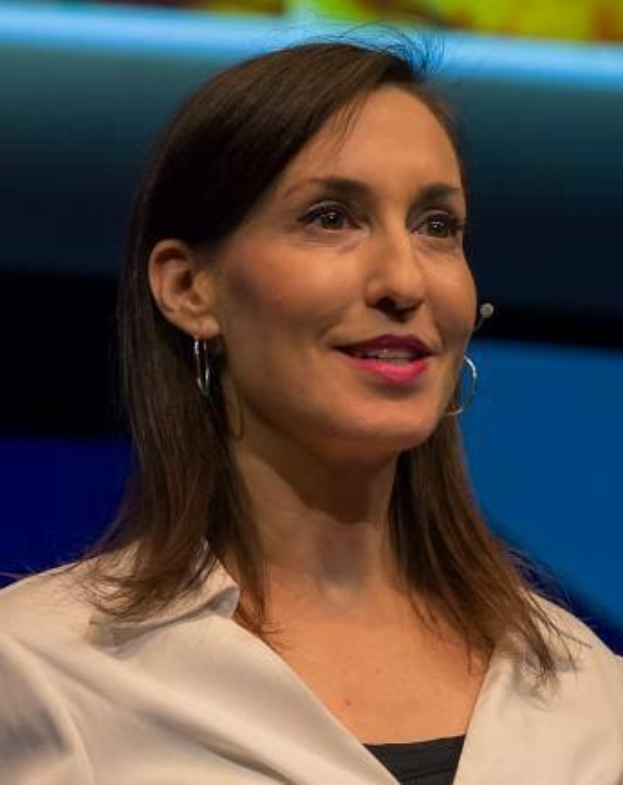 Eine Frau in einer weißen Jacke und einem schwarzen Oberteil sitzt vor einem blauen und gelben Hintergrund und trägt Ohrringe und ein Mikrofon während eines Interviews.