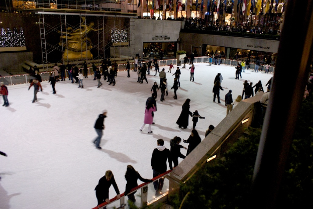 Eine Gruppe von Menschen, die auf einer vereisten Fläche umgeben von Gebäuden Eislaufen.
