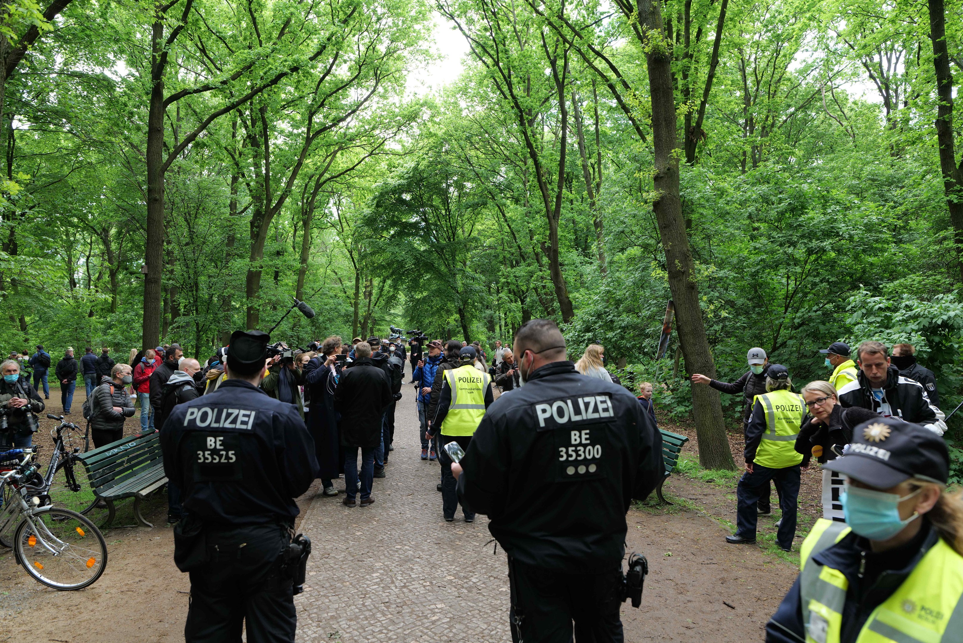 Eine Gruppe von Polizisten steht vor einer Menge bei einer Anti-Terror-Demonstration in Berlin, mit Fahrrädern und einer Bank im Vordergrund und Bäumen und Himmel im Hintergrund.