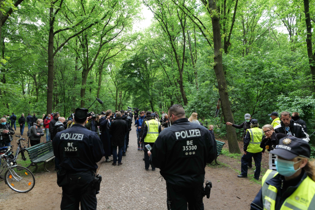 Eine Gruppe von Polizisten steht vor einer Menge bei einer Anti-Terror-Demonstration in Berlin, mit Fahrrädern und einer Bank im Vordergrund und Bäumen und Himmel im Hintergrund.