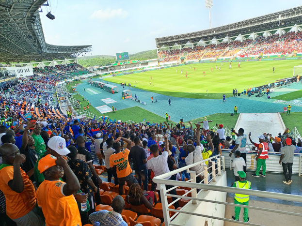 Eine große Menschenmenge in einem Stadion, die ein Fußballspiel ansieht, mit einem Schuppen mit Lichtern auf der linken Seite und Hügeln und einem klaren blauen Himmel im Hintergrund.