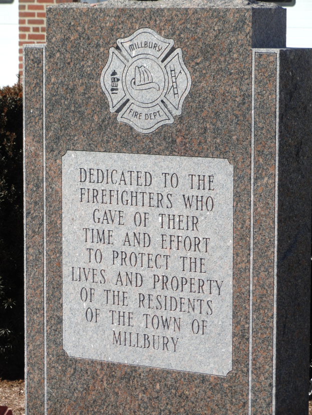 Gedenkstein mit einer Plakette für Feuerwehrleute in Millbury, vor einem Backsteingebäude mit umliegender Vegetation.