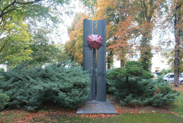 Große Metallskulptur in einem Park mit Bäumen, Pflanzen und zerrissenen Blättern, umgeben von Gebäuden unter einem klaren blauen Himmel.