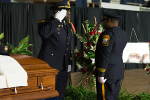 Zwei Polizeibeamte in Uniform stehen neben einem Sarg, der mit einem wei├čem Tuch bedeckt ist.