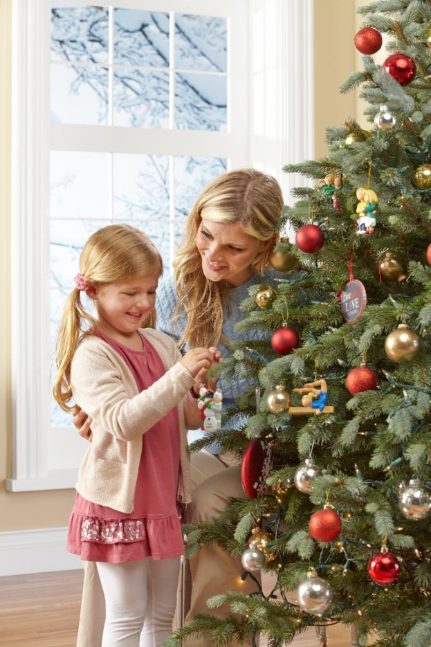 Ein geschmückter Weihnachtsbaum steht in einem Haus mit einer Frau und einem Kind im Vordergrund, einem Fenster und einer Wand im Hintergrund und Ästen vor dem Fenster.
