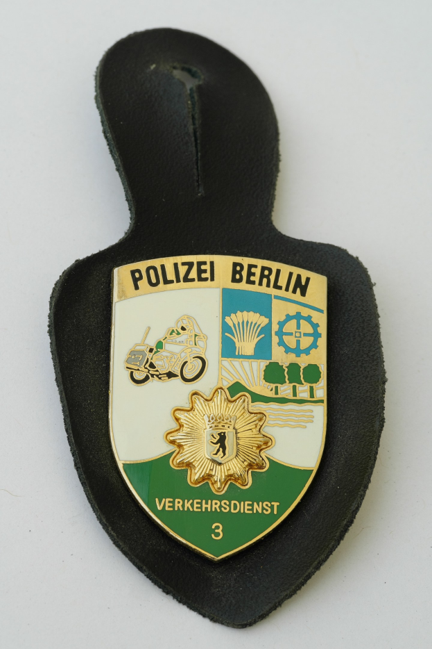 Ein schildförmiges Polizeiamblem mit der Aufschrift 'Polizei Berlin' auf einem blauen Hintergrund, einem weißen Rand und einem weißen Stern in der Mitte sowie schwarzer fetter Schrift auf einer Oberfläche.