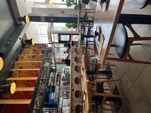 Ein Restaurantinnenraum mit Tischen und Stühlen in Reihen, Menschen, die stehen, Treppe auf der linken Seite, hängende Lampen und eine Pflanze im Hintergrund.