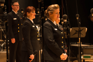 Eine Gruppe von Menschen, die vor Mikrofonen stehen, mit einem Mann und einer Frau, die Mikrofone halten, und Musikinstrumenten im Hintergrund.