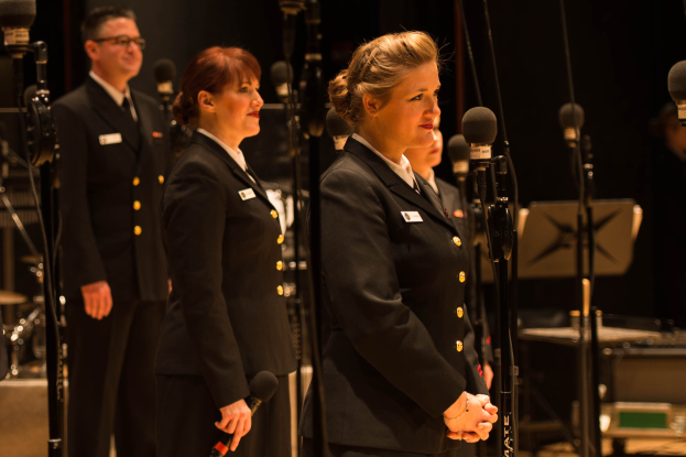 Eine Gruppe von Menschen, die vor Mikrofonen stehen, mit einem Mann und einer Frau, die Mikrofone halten, und Musikinstrumenten im Hintergrund.