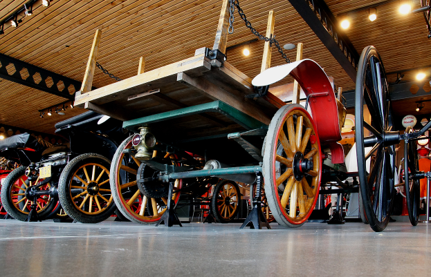 Ein altmodischer horse-drawn carriage in einem Museum ausgestellt, mit einer hölzernen Decke, die mit Lichtern und Ketten geschmückt ist, umgeben von anderen Fahrzeugen.