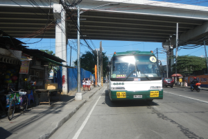 Ein grüner und weißer Bus fährt auf einer Straße neben einer Brücke, mit Strommasten und -kabeln entlang der Straße, Menschen, die gehen, Fahrräder, die geparkt sind, Geschäfte und Stände, die die Straße säumen, und Bäume mit einem klaren blauen Himmel im Hintergrund.