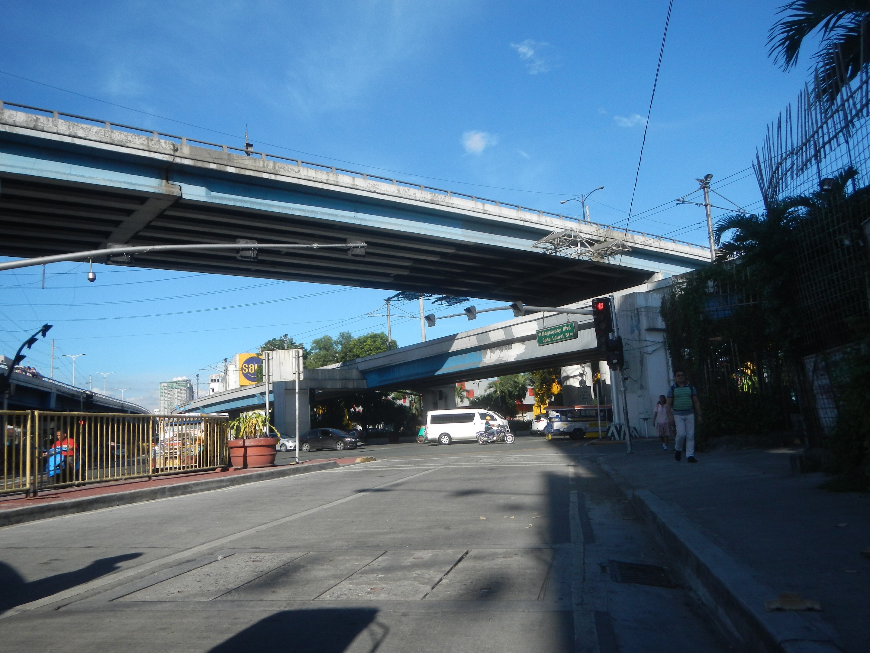 Stadtstraße mit einer Fußgängerbrücke darüber, Fahrzeuge auf der Straße, Fußgänger auf dem Gehweg, Beschilderung, Laternenpfähle, Bäume, Gebäude und ein bewölkter Himmel.