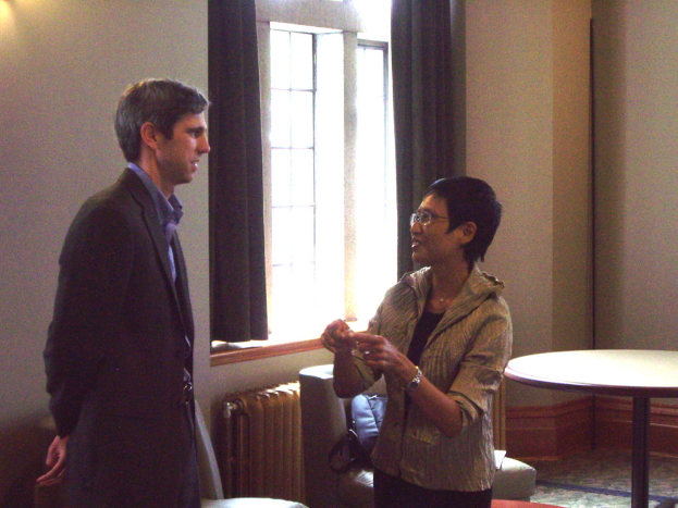 Eine Frau in einem Raum spricht zu einem Mann in einem schwarzen Anzug, der vor ihr steht, mit einem Tisch rechts und einer Tasche auf einem Stuhl hinter ihr.