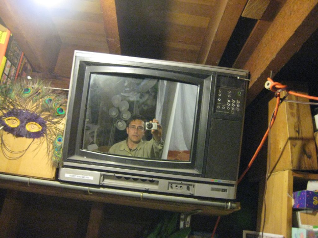 Ein Fernsehbildschirm mit einer Person, umgeben von Pfauenfedern, Büchern und Kartons.