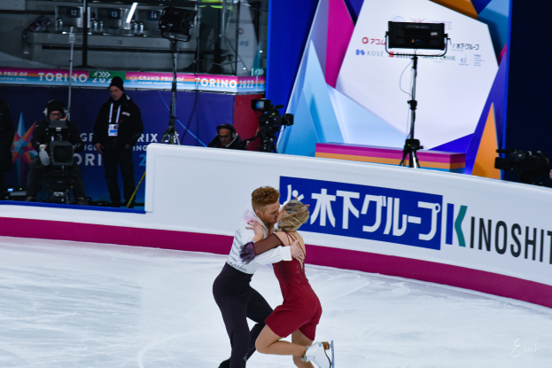 Ein Paar umarmt sich beim Eislaufen auf einer Eisbahn, im Hintergrund stehen einige Menschen, einige halten Kameras und eine Tafel mit Text, sowie Lichter und andere Gegenstände.