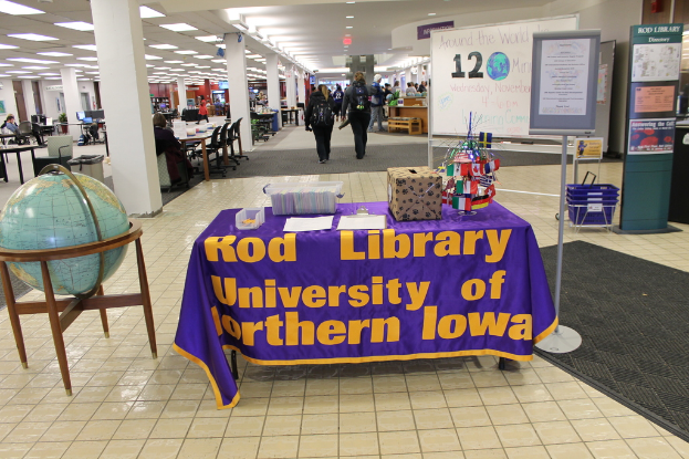 Eine Ausstellung mit einem Tisch, der verschiedene Landesflaggen, eine Box, Papiere und eine Fahne zeigt, eine Kugel links daneben, eine Mitteilungstafel rechts daneben mit einigen Leuten in der Nähe und mehreren Tischen und Stühlen im Hintergrund.