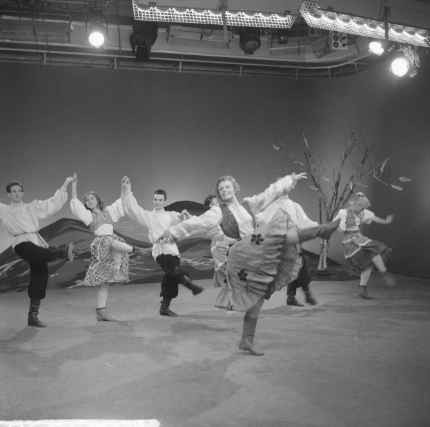 Eine Gruppe von Menschen, die auf einer Bühne tanzen, beleuchtet von Deckenlampen und von einer Wand umgeben, dargestellt in Schwarz-Weiß.
