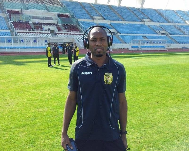 Ein Mann, wahrscheinlich der Trainer der ghanaischen Nationalmannschaft, steht auf einem Fussballfeld in der Nähe eines Stadions und trägt ein Headset sowie ein Objekt in der Hand, mit einigen Leuten im Hintergrund und dem Himmel über ihm.