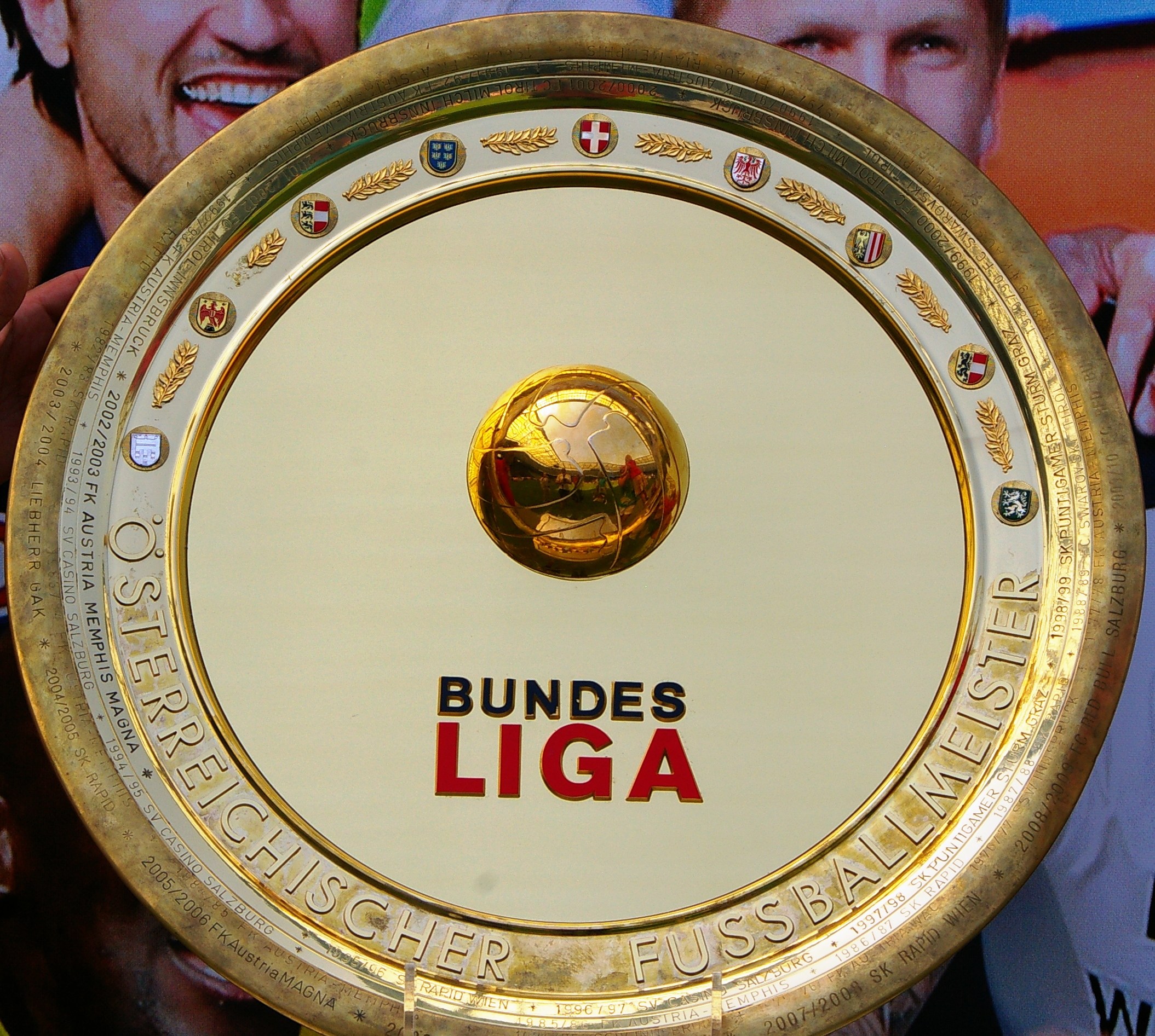 Goldener Schild mit 'Bundesliga'-Text in der Hand eines Mannes, im Hintergrund ein Banner mit drei Personen.