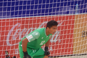 Ein Mann in grüner Torwartuniform kniet vor einem Tor mit einem Netz dahinter und einer Flasche rechts daneben.