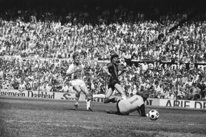 Eine Gruppe von Männern, die Fußball auf einem Feld spielen, mit Zuschauern und einem Banner im Hintergrund, das 'Bundesliga 1978-79 Ajax 1-1 Hannover 96' zeigt.