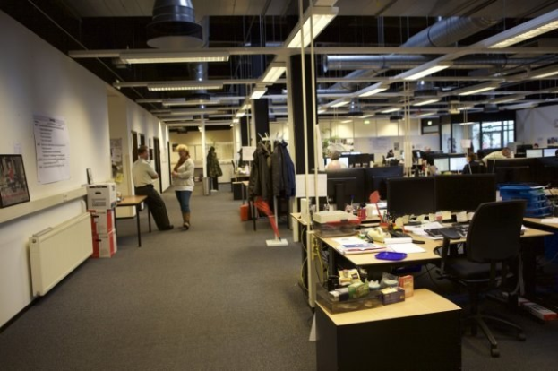 Großes Großraumbüro mit Menschen an Schreibtischen, Computern und Monitoren, umgeben von Papieren, Büchern, Kartons und an der Wand hängenden Tafeln mit Text und Bildern unter Deckenleuchten und Glasfenstern.