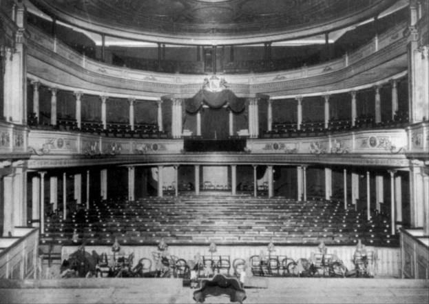 Schwarz-weiß-Foto des Theater Royal-Auditoriums in London, das Stühle, Treppen, Geländer, Säulen, Lampen und einen Kronleuchter mit einigen sitzenden und stehenden Menschen zeigt.