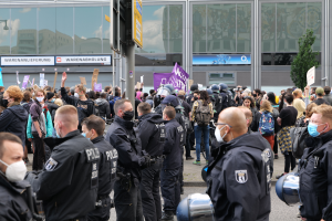Eine große Gruppe von Menschen steht vor einem Gebäude, einige halten Schilder und tragen Helme, mit einem Mast mit einer Tafel im Vordergrund und einem Baum im Hintergrund, scheinbar protestierend.
