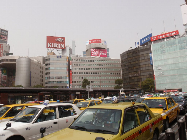 Eine Stadtansicht mit Fahrzeugen auf der Stra├če, Geb├Ąuden, B├Ąumen, Masten mit Schildern, und Text auf den Schildern, einem Zug im Hintergrund und einem Gel├Ąnder unten rechts.