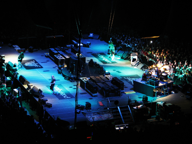 Eine Gruppe von Menschen, die auf einer Bühne mit Lautsprechern, Scheinwerfern und Mikrofonen performs, umgeben von einer großen Menge in einem dunklen Hintergrund.