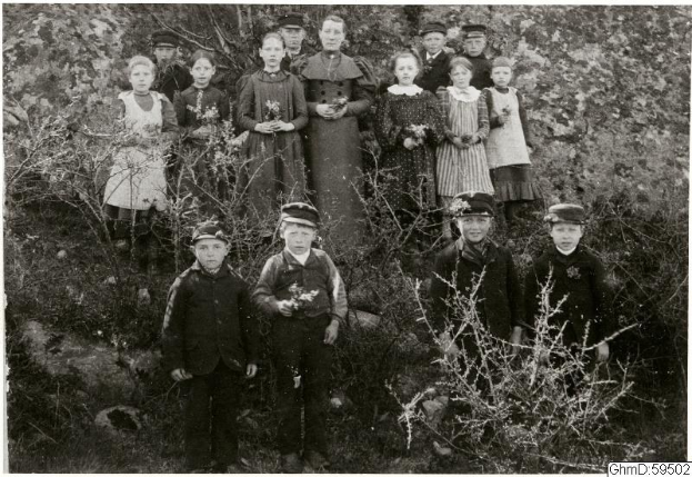 Schwarz-weiß-Foto einer Gruppe von Kindern in Mützen, einige halten Blumen, stehen in einem Garten mit Pflanzen und Bäumen im Hintergrund, beschriftet mit "1930er-Kinder im Garten."