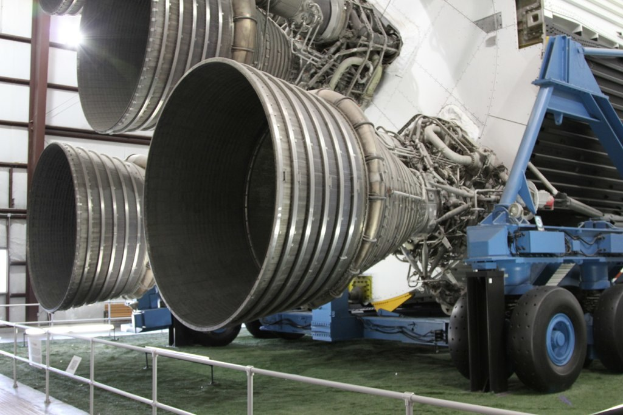 Ein großes metallenes Fahrzeug mit Rädern und einer Stange ist auf einem grasbewachsenen Bereich geparkt, mit einem Zaun und einer Wand mit Metallstangen im Hintergrund.