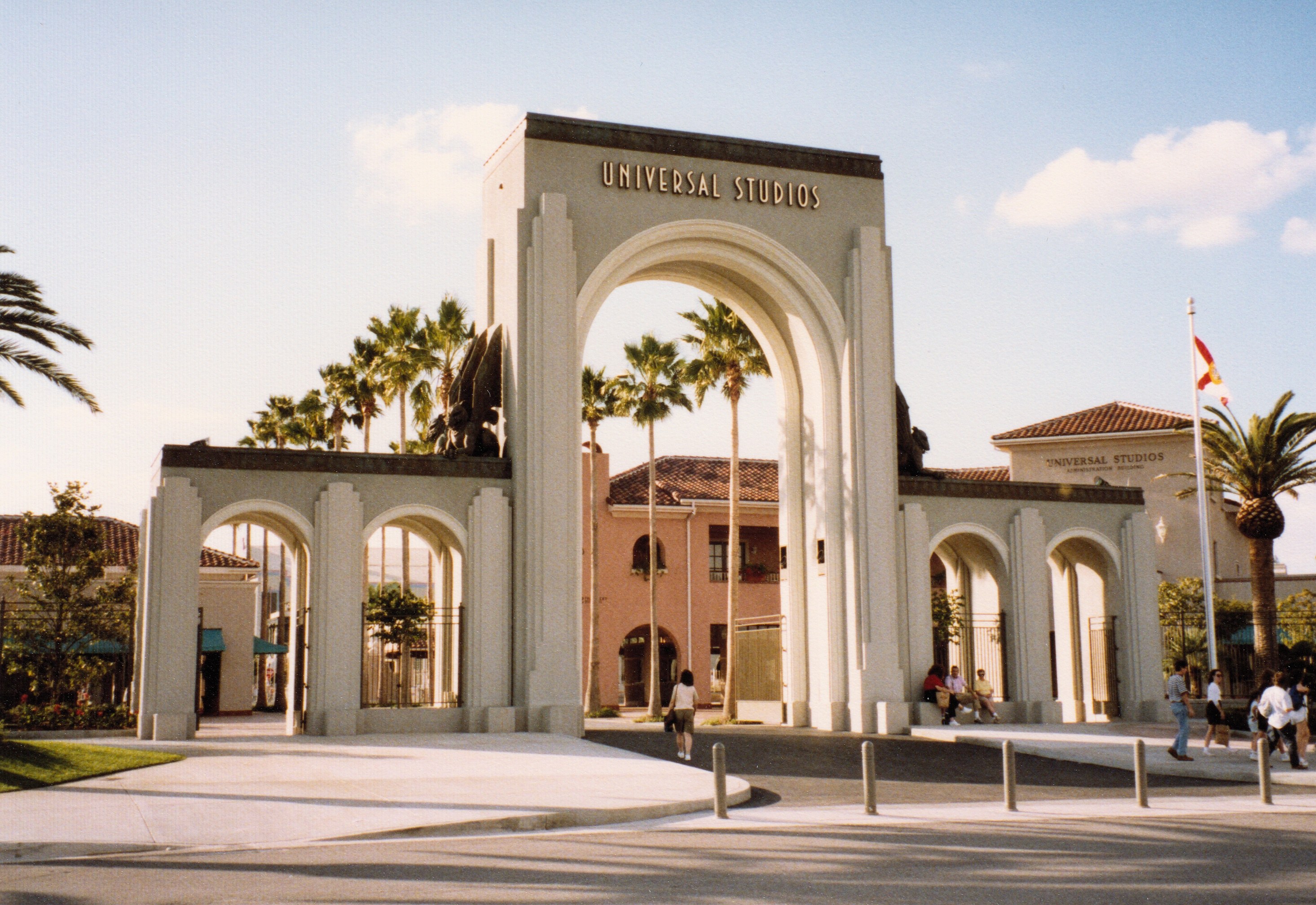 Eingang zu Universal Studios Hollywood mit einem Bogen, einer Statue, Menschen, Bäumen, Gebäuden, einem Fahnenmast mit einer Flagge und einem bewölkten Himmel.