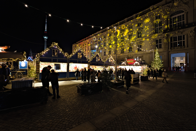 Ein lebendiger Weihnachtsmarkt in Berlin, Deutschland, mit Menschen um hell erleuchtete Stände, festliche Dekorationen und Gebäude mit Fenstern im Hintergrund unter einem dunklen Himmel.