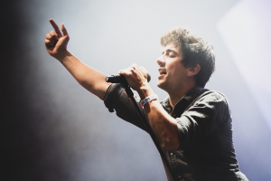 Ein Mann in schwarzem Hemd und Jeans singt leidenschaftlich in ein Mikrofon auf einer hell erleuchteten Bühne, mit geschlossenen Augen und stylischem Haar, während das Publikum im Hintergrund applaudiert.