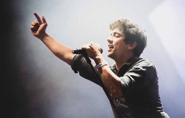 Ein Mann in schwarzem Hemd und Jeans singt leidenschaftlich in ein Mikrofon auf einer hell erleuchteten Bühne, mit geschlossenen Augen und stylischem Haar, während das Publikum im Hintergrund applaudiert.