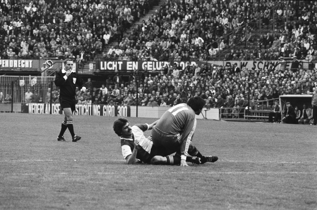 Ein Schwarz-Weiß-Foto von zwei Fußballspielern, wobei einer den anderen umgrätscht, mit Zuschauern und Stadionstrukturen im Hintergrund und einer Person mit einem Gegenstand auf der linken Seite.
