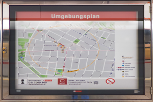 Eine Bushaltestelle mit einem großen Bildschirm, der eine Karte von München, Deutschland, zeigt, eingerahmt von einem Metallrahmen und einem unscharfen Hintergrund.