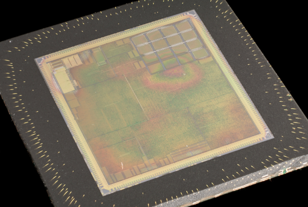 Nahaufnahme eines rechteckigen Halbleiterchips mit metallischem Glanz, umgeben von einem dünnen Metallrahmen und kleinen Löchern entlang seiner leicht erhöhten Kanten auf schwarzem Hintergrund.